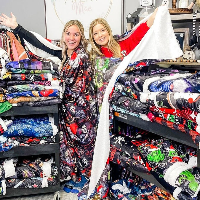 Two people standing behind a lot of large, colorful, Sherpa blankets in a retail setting with clothing racks in the background.