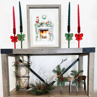 Winter home decor and holiday accents, with red and green taper candles, a framed fireplace print, a JOY wood sign, evergreen and a wood reindeer.