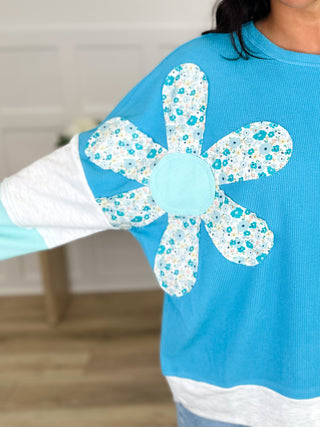 Long-sleeve cotton-blend top with large blue floral four-petal applique on teal body