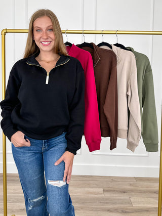 A woman stands in front of a gold clothing rack wearing a black half-zip sweatshirt and blue jeans. Five other sweatshirt colors are hanging on the rack behind her: black, bright pink, brown, beige, and olive green.