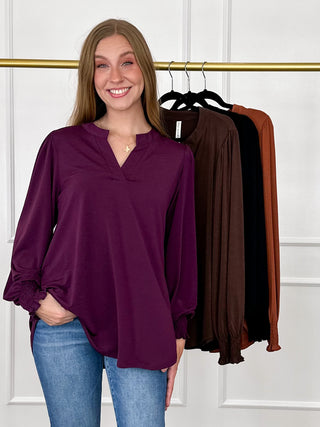 A woman wearing a long-sleeve henley-style top in deep plum purple, standing in front of a clothing rack with three additional top colors visible: brown, black, and rust orange.