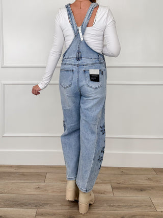 Medium-wash denim bib overalls with adjustable back straps, front pockets, floral knee embroidery