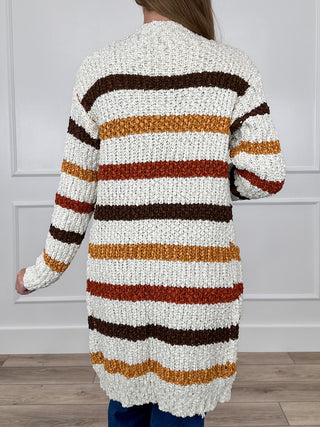 Back view: ivory long knit cardigan with long sleeves and bold brown, orange, black stripes.