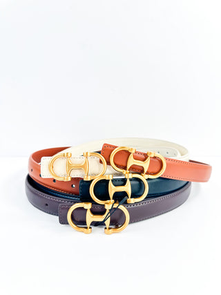 Leather belt stack in black, brown, and bone white with gold-tone buckle hardware.