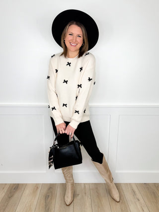 Women's outfit: white sweater with black bow-star print, black pants, beige boots, black handbag.