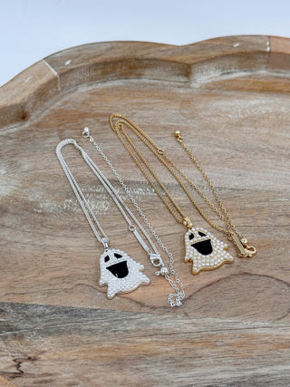 Two ghost-shaped necklace pendants with black mouths are displayed on a rustic wooden tray. Two pendants are silver-toned with clear rhinestones, and one is gold-toned with white rhinestones.