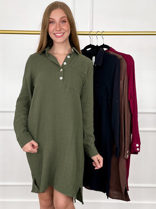 A woman wearing an olive green long-sleeve button-front shirt dress stands in front of a clothing rack displaying other shirt dresses in black, brown, and burgundy.