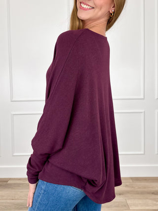 A woman wearing a loose, long-sleeve knit top in deep plum purple, paired with blue jeans, shown from a side-back angle against a white paneled wall.