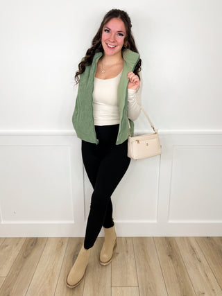 Women’s outfit with green jacket, cream top, black pants, beige boots, handbag