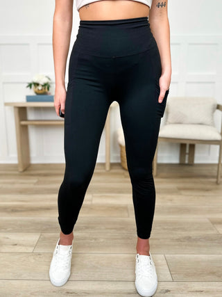 A woman wearing black pocket leggings and white sneakers in a room with wooden flooring and a white wall.