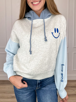 Cropped graphic hoodie with heather grey body, blue contrast sleeves, and smiley face emblem