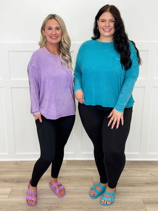 Two women wearing long-sleeve tops and black leggings standing against a white wall.