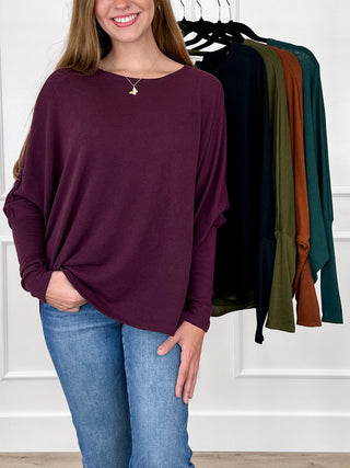 A woman wearing a loose, long-sleeve boat-neck top in deep burgundy with blue jeans, standing in front of a white paneled wall. A rack of other long-sleeve tops in black, green, olive, and rust is visible behind her.