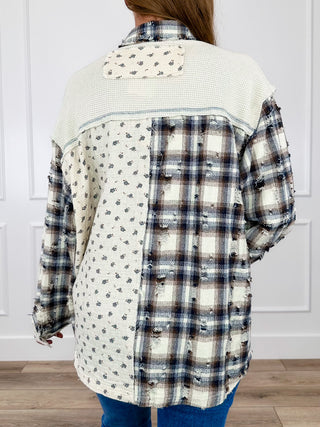 A person shown from the back wearing a patchwork button-up shirt with mixed fabric panels: one side is cream with small dark floral print, the other side is a dark plaid pattern, and the upper yoke is cream waffle-knit. The person is also wearing blue jeans, standing indoors against a white paneled wall.
