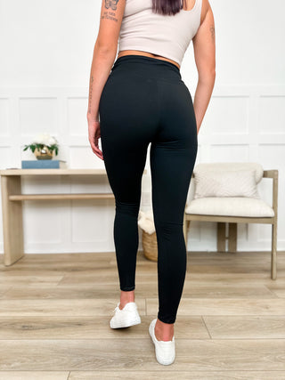 Person wearing black leggings and a beige top in a room with wooden flooring and a white wall.