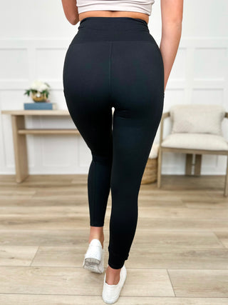 A woman wearing black pocket leggings and white sneakers in a room with wooden flooring and a white wall.