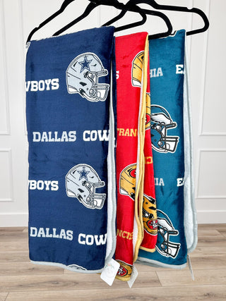 Team-logo sherpa throw blankets in blue, red, and teal hanging on hooks