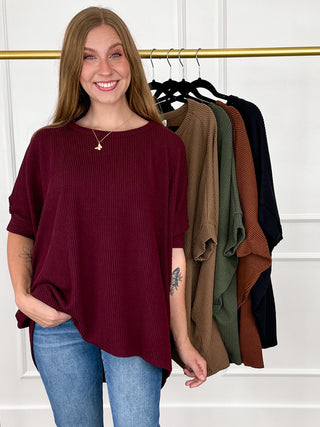 A smiling woman stands indoors wearing a loose ribbed short-sleeve top in deep burgundy with blue jeans. Behind her, a gold clothing rack holds several other ribbed tops in brown, olive green, rust orange, and black.
