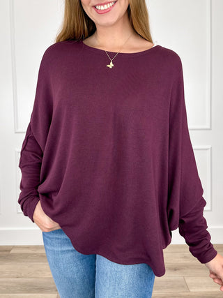 A woman wearing a loose, long-sleeve boat-neck top in deep plum purple, paired with blue jeans, standing indoors against a white paneled wall.