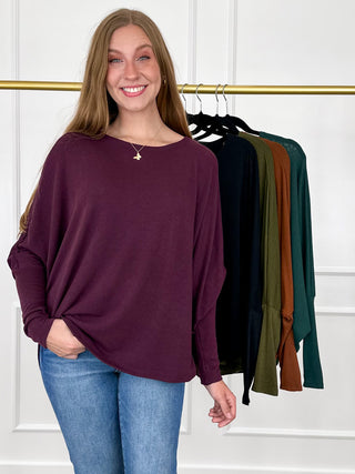 A woman wearing a loose, long-sleeve boat-neck top in deep plum purple, paired with blue jeans. She is standing in front of a clothing rack displaying other color options (black, olive green, brown, teal) against a white paneled wall.