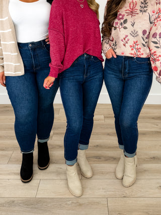 Three models wearing Judy Blue dark blue skinny jeans, styled with different tops and ankle boots, standing on a light wood floor.