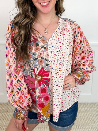 A woman wearing a long-sleeve split-pattern blouse with one side featuring a pink floral print and the other side featuring a small red floral print on a cream background, paired with denim shorts.