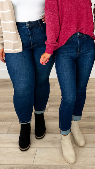 Two models standing side-by-side wearing dark wash Judy Blue skinny jeans, with one in black ankle boots and the other in cream ankle boots, standing on a light wood floor.