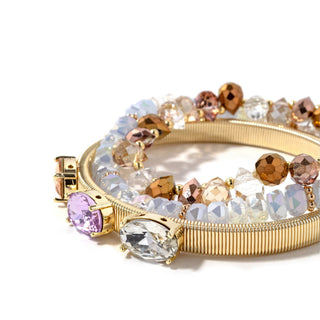 Close-up gold-tone bracelet stack with clear stones, brown beads, and purple charm