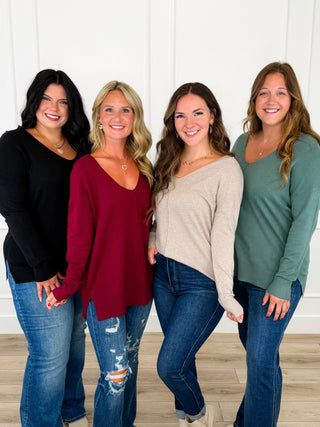Four women standing together in a bright indoor setting, each wearing a different long-sleeve top in black, burgundy, beige, and sage green, paired with blue jeans.