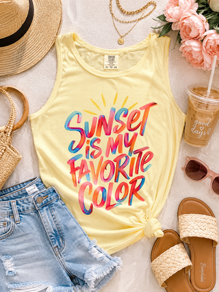 Yellow tank top with 'Sunset is my favorite color' text, in a styled flat-lay with denim shorts, sandals, and flowers on a light surface.