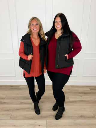 Black quilted puffer vest layered over a rust and a wine knit sweater with black leggings and black booties.