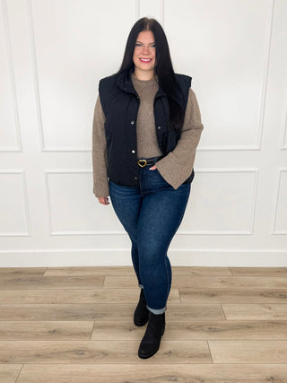 Brown speckled knit sweater layered under a black quilted vest with dark wash skinny jeans and black booties.