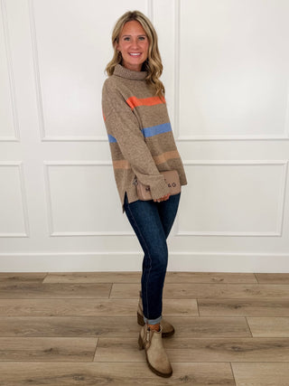 Taupe striped turtleneck sweater with orange/blue bands and dark blue jeans