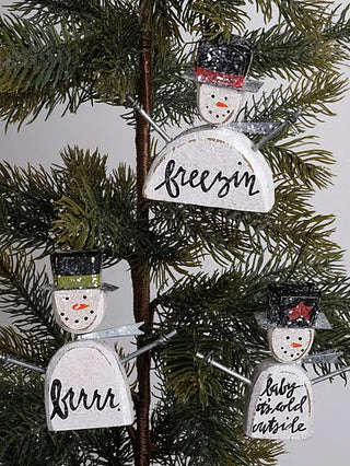 Three wooden snowman ornaments hanging on branches: 'freezin', 'brrr', 'baby inside'