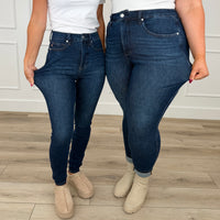 Two models wearing dark wash high-waisted Judy Blue skinny jeans, stretching the fabric at the hips to demonstrate the stretchiness, standing on a light wood floor.