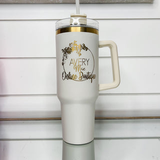 Cream gold-accent tumbler with clear lid, side handle, and front logo.