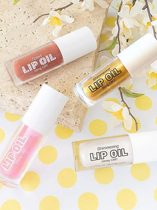 Assorted tinted and shimmering CBD lip oil bottles in pink, gold, and brown shades.