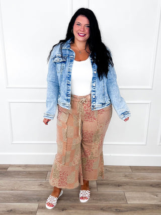 Light wash denim jacket layered over a white tank with tan floral patchwork wide leg pants and white sandals.