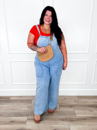 Light wash denim jumpsuit with tie neck straps styled over a red top with a tan woven crossbody bag.