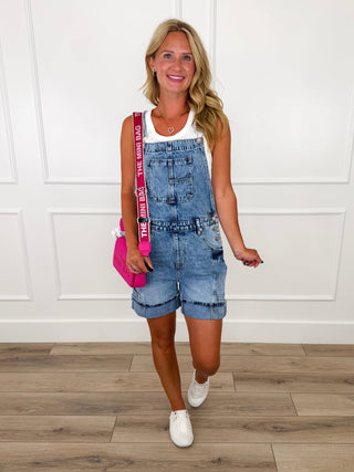 Woman wearing medium wash denim overall shorts over a white ribbed tank with white sneakers and a pink crossbody bag.